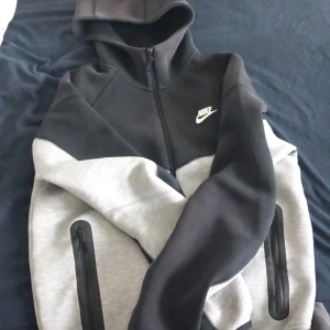 Svart och grå hoodie från Nike - Säljer en snygg svart och grå hoodie från Nike med dragkedja framtill och två stora fickor med dragkedjor. Jackan har huva och klassisk Nike-logga på bröstet. Perfekt för en sportig och avslappnad stil. Den e knappt använd. Pris kan diskuteras