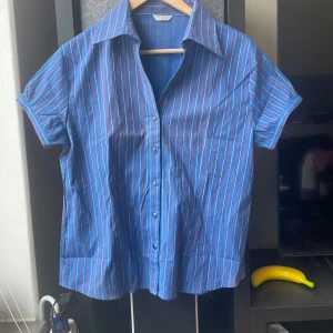 Blå kortärmad skjorta med ränder från A&W - Säljer en blå skjorta med röda och vita ränder från A&W. Skjortan har korta ärmar, klassisk krage och knappar framtill. Perfekt för dig som gillar retrostil och vill sticka ut lite extra.