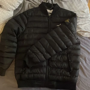 Svart pufferjacka från Stone Island - Säljer en svart pufferjacka från Stone Island med klassisk logotyp på ärmen. Jackan har dragkedja framtill, två sidofickor med dragkedja och ribbade muddar. Perfekt för kyligare dagar och riktigt snygg streetstil. (AA)