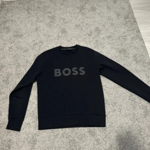Svart sweatshirt från BOSS - Säljer en svart sweatshirt från BOSS med rund halsringning och tryckt logga framtill. Tröjan har långa ärmar och en klassisk passform, perfekt för en stilren och enkel look.