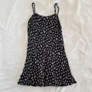 Svart blommig klänning från Indiska - Supersöt svart klänning från Indiska med små vita och lila blommor. Klänningen har smala axelband och är ärmlös, perfekt för varma dagar. Den har en lätt utställd passform och känns luftig och skön. Den är lite trasig uppe vid ens axelbandet (sista bilden)