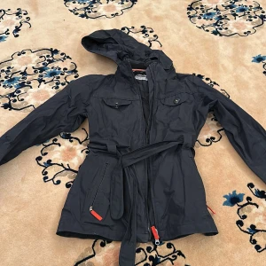 Svart vindjacka med huva och bälte - Säljer en svart vindjacka med huva och bälte i midjan. Jackan har dragkedja framtill, fickor med lock och knappar samt praktiska detaljer som gör den perfekt för blåsiga dagar. Snygg och funktionell design med justerbar passform.