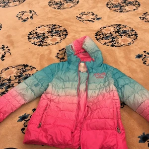 Färgglad pufferjacka/dunjacka med huva - Säljer en pufferjacka i snygg ombre-design med blått upptill och rosa nedtill. Jackan har huva, dragkedja och två fickor framtill. Perfekt för kyliga dagar och ger en cool look. 