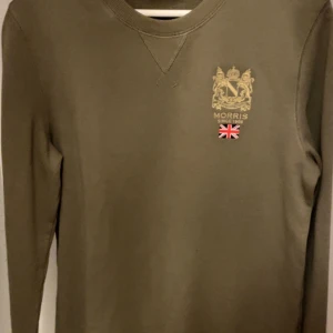 Olivgrön sweatshirt från Morris - Säljer en olivgrön sweatshirt från Morris med guldfärgat broderat emblem och liten brittisk flagga på bröstet. Tröjan har rund halsringning och långa ärmar. Perfekt för en stilren och avslappnad look.