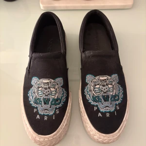 Svarta slip-on sneakers från Kenzo - Svarta slip-on sneakers från Kenzo med broderad tiger och texten 'Paris' framtill. Skorna har vit platt sula och är tillverkade i textil med Kenzo-logga bak på hälen.