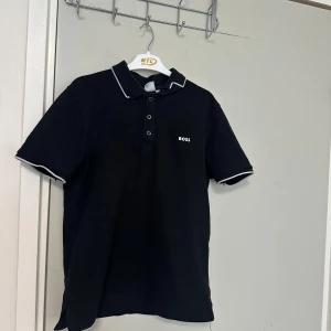 Svart pikétröja från BOSS - Säljer en svart pikétröja från BOSS med vit kant på krage och ärmslut. Tröjan har klassisk pikékrage, knappar framtill och liten BOSS-logga på bröstet. Perfekt för en stilren och sportig look.