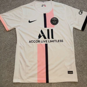 PSG fotbollströja 2021/22 från Nike - Säljer en vit PSG fotbollströja från Nike med svart och rosa vertikal rand på framsidan. Tröjan har korta ärmar, svart Nike-logga och PSG-märke på bröstet samt sponsortryck. Materialet är lätt och andas, perfekt för match eller träning.