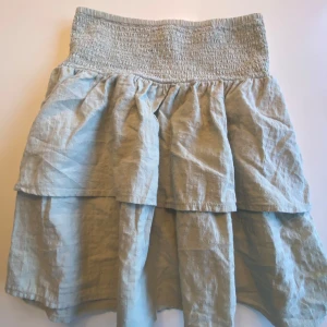 Beige volangkjol från Kappahl - Säljer en beige kjol från Kappahl med bred smock i midjan och dubbla volanger. Kjolen är luftig och har en avslappnad passform, perfekt för varmare dagar. Tillverkad i ett lätt material som känns skönt mot huden.