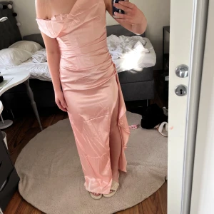 Ljusrosa långklänning från Babyboo - Säljer en elegant ljusrosa långklänning från Babyboo med offshoulder-skärning och draperad detalj vid bysten. Klänningen har en hög slits framtill och är tillverkad i ett glansigt satinliknande material. Perfekt för speciella tillfällen! Jag säljer klänningen för att jag beställde samma klänning men i två storlekar. Klänningen gick inte att lämna tillbaka även fast att den är helt oanvänd och har lapparna kvar. Den är från babyboo och där kunde man inte returnera varorna. Original förpackning