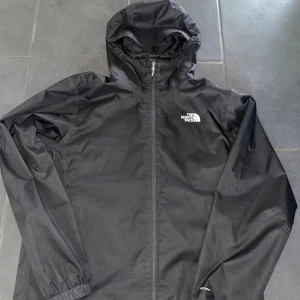 Svart vind/regn jacka från The North Face - Säljer en svart vind/regn jacka från The North Face med huva och dragkedja framtill. Jackan har lång ärm, elastiska muddar och meshfoder på insidan. Perfekt för blåsiga/regniga dagar och enkel att packa ner i väskan.