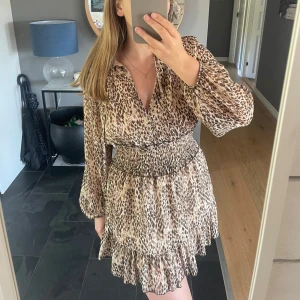 Leopardmönstrad klänning från Zara - Säljer en snygg leopardmönstrad klänning från Zara med långa ärmar och smockad midja. Klänningen har v-ringning och volangdetaljer nertill, vilket ger en trendig och feminin look. Perfekt för dig som vill sticka ut!