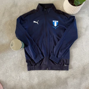 Mörkblå Malmö FF träningsjacka från Puma - Säljer en mörkblå träningströja från Puma med Malmö FF-logga på bröstet. Tröjan har dragkedja framtill, långa ärmar och resår vid ärmslut och midja. Perfekt för fotbollsträning eller som supporterplagg.Den är lite urtvättad och nopprig men på insidan är den väldigt skön och bra för träning