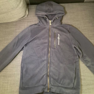  hoodie från Sail Racing - Mörkgrå hoodie från Sail Racing med dragkedja framtill och huva. Tröjan har en bröstficka med dragkedja och logga tryckt på vänster sida. Perfekt för en avslappnad stil.