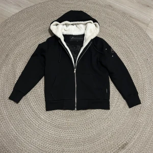 mose knuckels hoodie/jacka - Svart hoodie från Moose Knuckles med dragkedja framtill och vit fuskpäls i huvan. Jackan har två fickor fram och ribbade muddar vid ärmslut och nederkant. Perfekt för kyliga dagar när du vill vara både varm och snygg.
