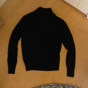 Svart stickad polotröja med dragkedja från Uniqlo - Säljer en svart stickad polotröja från Uniqlo med dragkedja vid halsen. Tröjan har lång ärm och ribbade muddar vid ärmslut och nederkant. Perfekt för lager-på-lager under kyliga dagar, men även för en sen sommarkväll!