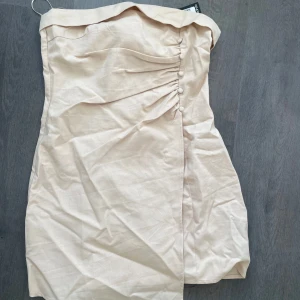 Champagnefärgad axelbandslös klänning från Boohoo - Säljer en champagnefärgad axelbandslös kortklänning från Boohoo med dekorativa knappar och drapering framtill. Klänningen har en figurnära passform och dragkedja i ryggen. Aldrig använd