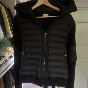 Svart moncler cardigan  - Svart moncler cardigan  från Moncler med stickade ärmar och en huva. Jackan har dragkedja framtill, fickor med tryckknappar och en extra ficka med dragkedja på ärmen. Kombinerar quiltad polyester med stickade detaljer för en riktigt snygg look. Perfekt för sommarkvällar. Storlek M passar S 