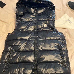Svart dunväst från Moncler - Säljer en svart, glansig dunväst från Moncler med huva och dragkedja framtill. Västen har två fickor med dragkedja och klassisk Moncler-logga på bröstet. Perfekt för lager-på-lager och riktigt snygg till streetwear-stilen. Sitter helt ok på mig som är 170 lite stor