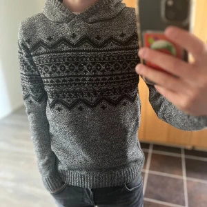 Stickad hoodie - Säljer en grå stickad hoodie med svart mönster över bröstet och armarna. Tröjan har huva, långa ärmar och ribbade muddar vid ärmslut och nederkant. Perfekt för kalla dagar och riktigt snygg till jeans. Passar S