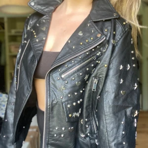 Svart skinnjacka med nitar och stjärnor - Riktigt snygg svart skinnjacka med coola detaljer som silverfärgade stjärnor, hjärtan och nitar över hela jackan. Jackan har dragkedjor, bred krage och en edgy bikerstil. Perfekt för dig som vill sticka ut och addera attityd till din outfit.