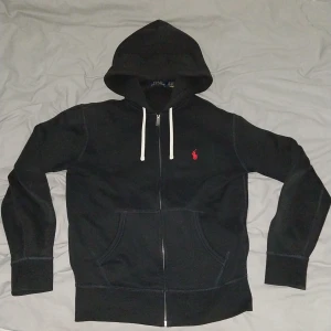 Svart hoodie från Polo Ralph Lauren XS - Svart hoodie från Polo Ralph Lauren i storlek XS. Klassisk design med vit snörning i huvan, dragkedja framtill och röd broderad logga på bröstet. Mjuk bomullskänsla och ficka framtill. Perfekt för en chill och stilren look.