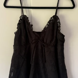 Blus  - Supersöt svart blus med smala axelband, volangdetaljer vid urringningen och en liten rosett framtill. Toppen har ett diskret blommönster i tyget och v-ringad hals. Perfekt för dig som gillar romantiska detaljer och en feminin look.