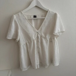 Vit blus med v-ringning och kort ärm - Söt vit blus från shein med v-ringning och korta ärmar. Blusen har små broderade prickar i tyget och en lätt volang under bysten som ger en luftig känsla. Perfekt för vår och sommar, enkel att matcha till jeans eller kjol.