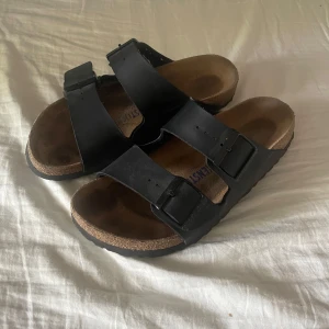 Svarta Birkenstock sandaler i skinn - Klassiska svarta Birkenstock sandaler med två breda remmar i skinn och justerbara spännen. Fotbädden är i kork och sulan är platt och bekväm. Perfekta för sommardagar och enkel att matcha med allt. Stora i storleken och passar nog 38-40.