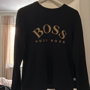 Svart sweatshirt från Hugo Boss - Svart sweatshirt från Hugo Boss med stort BOSS-tryck i guld på bröstet. Tröjan har rund hals, långa ärmar och ribbade muddar vid ärmslut och nederkant. Perfekt för en clean och stilren look med exklusiv känsla.