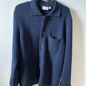 Mörkblå stickad overshirt från Kappahl - Snygg mörkblå stickad overshirt från Kappahl i storlek S. Skjortan har klassisk krage, knäppning framtill med stora knappar och en bröstficka. Perfekt att slänga över en t-shirt för en chill look.