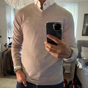 Vit v-ringad tröja Tiger of Sweden - Snygg vit v-ringad tröja från Tiger of Sweden i mjuk bomullsblandning med diskret broderad logga på bröstet. Klassisk passform och ribbade muddar, perfekt att bära över skjorta för en clean och stilren look.