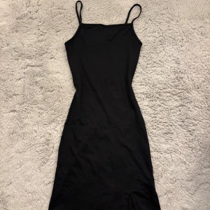 Svart tight klänning med smala band - En svart, figurnära klänning från Gina Tricot med smala axelband och rak halsringning. Klänningen är enkel och stilren, perfekt för dig som gillar minimalistisk stil. Materialet känns mjukt och stretchigt, vilket gör den bekväm att bära.