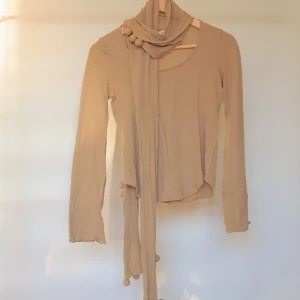 3.1 Phillip Lim top - Supersnygg beige långärmad topp från 3.1 Phillip Lim. Unik scarf-detalj med bollar som hänger ner framtill, perfekt för att ge outfiten en cool twist. Rund halsringning och figurnära passform. Passar dig som gillar stilrena men annorlunda plagg.