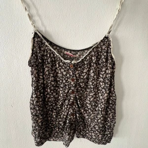 Blommigt linne från Brandy Melville - Supersöt linnetopp med småblommigt mönster i svart, beige och vitt. Tunt och luftigt material, knappar framtill och smala axelband med spetskant. Lite slitet skick, axelbanden känns lite torra och uttöjda och det har två små hål i ryggen, det är dock inget som syns när linnet sitter på.