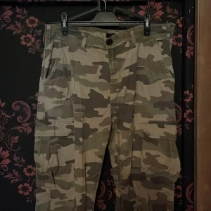 Cargobyxor i camo från Saligia - Cargobyxor i klassiskt camouflage-mönster från Saligia. Byxorna har flera stora fickor på sidorna och en loose passform. Perfekta för dig som gillar streetstyle och vill ha en avslappnad look.