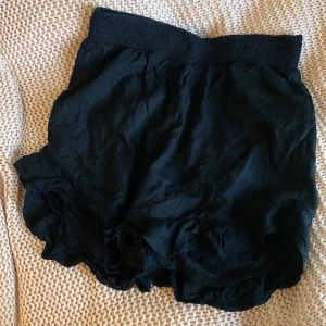 Svart volangkjol/shorts med resårmidja - Svarta shorts/kjol med bred resår i midjan och volangkant nertill. Lätta och luftiga, perfekta för varma dagar. Lätt och luftigt material i storlek 146/152.