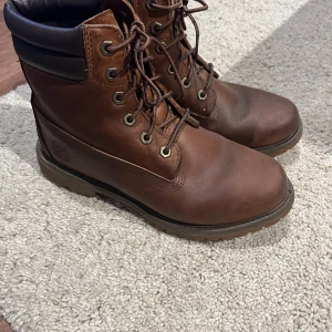 Bruna Timberland boots i skinn - Klassiska bruna boots från Timberland i robust skinn med snörning och vadderad svart kant upptill. Grov sula för extra grepp och slitstyrka. Perfekta för höst och vinter när du vill ha både stil och funktion och vattentäta. Strl 39,5