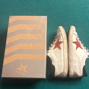 Golden Goose Superstar sneakers vit/röd - Golden Goose Superstar sneakers i vitt skinn med ikonisk röd stjärna på sidan och svarta detaljer vid hälen. Klassisk låg modell med grå snörning och platt sula. Snyggt slitna för en cool, avslappnad look. Kommer med originalkartong. De är ganska slitna men det är därför priset är billigt. Priset är ej hugget i sten