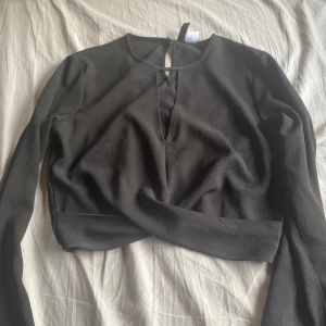 Svart långärmad topp från H&M Divided - Svart långärmad topp från H&M Divided med croppad passform. Toppen har en djup v-ringning framtill och en liten knapp i nacken. Tillverkad i ett lätt strukturerat polyestertyg som ger en snygg textur. Perfekt till jeans eller kjol för en clean look.