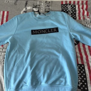 Ljusblå Moncler sweatshirt i bomull - Snygg ljusblå sweatshirt från Moncler med klassisk rund hals och stort Moncler-tryck på bröstet. Tröjan är i mjuk bomull och har långa ärmar samt ribbade muddar vid ärmslut och nederkant. Perfekt för dig som gillar stilrena och exklusiva plagg.