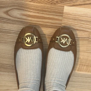 ballerinaskor från Michael Kors - Snygga bruna ballerinaskor från Michael Kors i skinn med guldigt MK-spänne framtill. Skorna har rund tå, perfekt för en klassisk look. 