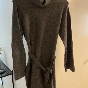 Mörkgrå stickad klänning med polokrage - Mysig mörkgrå stickad klänning med polokrage och knytskärp i midjan. Långärmad modell med ribbade muddar och avslut. Perfekt för kalla dagar när du vill vara både bekväm och snygg.