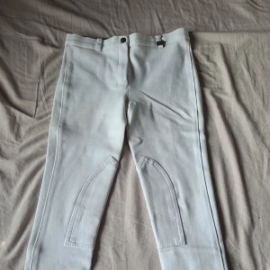 Vita ridbyxor CRW stl 146/152 - Snygga vita ridbyxor från CRW i storlek 146/152. Byxorna har förstärkta knäpartier, hällor för bälte och stängs med knapp och dragkedja. Tillverkade i bomull med lite stretch för skön passform. Perfekta för ridning!