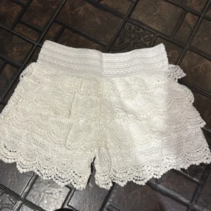 Vita spetsiga shorts med volang - Superfina vita shorts med spets och volangkant. De har hög midja och ett elastiskt midjeband som sitter bekvämt. Perfekta för sommardagar när du vill ha en söt och luftig look. Spetsen ger shortsen en romantisk vibe och de är lätta att matcha med crop tops eller linnen.