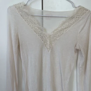 Vit långärmad topp med spets från Intimissimi - Superfin vit långärmad topp från Intimissimi med v-ringning och spetsdetaljer längs halsringningen. Tunn och mjuk i bomullsmix, Den har två små hål men dem syns inte så mycket när man har på sig den.