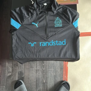Svart Marseille half zip tröja Puma  - Svart tränings-tröja från Puma med Olympique de Marseille-logga och blå detaljer. Half zip framtill, långärmad och tillverkad i polyester. Perfekt för fotboll eller chill. Randstad-tryck på bröstet och klassisk Puma-logga i blått.