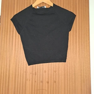Svart croppad topp från Zara - En stilren svart croppad topp från Zara med korta ärmar och rund hals. Toppen är ribbad och har en tight passform som sitter snyggt på kroppen. Perfekt att matcha med höga jeans eller kjol för en clean look.