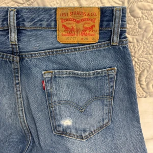 Levi's 501 blå jeans med slitningar - Klassiska Levi's 501 jeans i ljusblå tvätt med raka ben och slitningar på knäet och bakfickan. Jeansen har hög midja, knappgylf och den ikoniska läderpatchen bak. Perfekta för en avslappnad och trendig look. Vintage jeans! Pris går att diskuteras.