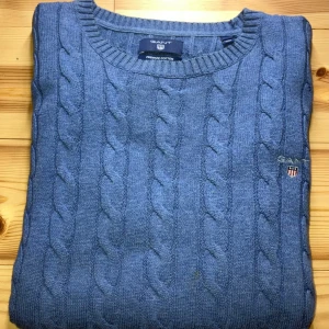 Blå kabelstickad tröja från GANT - Snygg blå kabelstickad tröja från GANT i 100% bomull. Klassisk rund halsringning och broderad GANT-logga med sköld på bröstet. Perfekt för dig som gillar en stilren och tidlös look. Passar lika bra till jeans som till chinos.