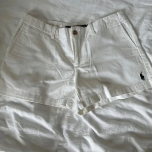 Vita bomullsshorts från Ralph Lauren - Snygga vita shorts från Ralph Lauren Sport i 100% bomull. Klassisk design med raka ben, bälteshällor, knäppning fram och två bakfickor med knappar. Broderad svart logga på ena benet ger en clean och sportig vibe. Perfekta för varma dagar. Shortsen är i storlek US4